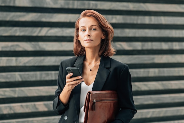 Zdjęcie bizneswoman w średnim wieku trzymająca skórzaną teczkę i smartfon