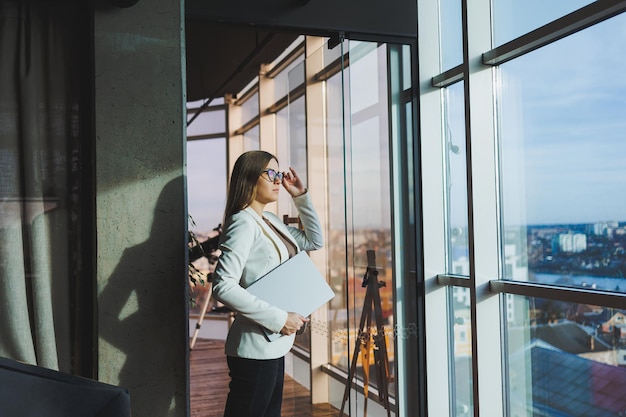 Bizneswoman z długimi włosami w białej kurtce i okularach stoi z laptopem w dłoniach w przestrzeni biurowej przy oknie i patrzy na miasto Pracuj w nowoczesnym biurze z dużymi oknami
