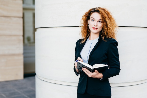 Bizneswoman z kręconymi włosami, pomalowanymi na czerwono ustami, ubrany w eleganckie ubrania, pisze w swojej książeczce