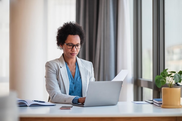 Bizneswoman z papierami pracuje na laptopie siedząc przy biurku