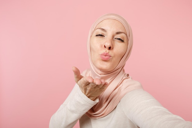 Bliska arabskiej kobiety muzułmańskiej w hidżab lekkie ubrania pozowanie na białym tle na różowym tle. Koncepcja życia religijnego ludzi. Makieta miejsca na kopię. Robienie selfie na telefonie komórkowym, wysyłanie pocałunku w powietrzu.