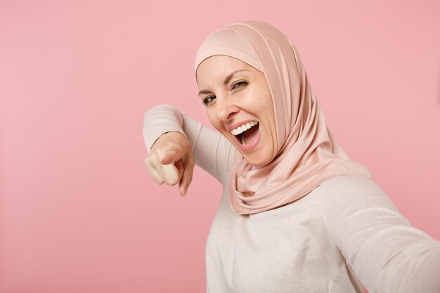 Bliska arabskiej muzułmańskiej dziewczyny w lekkie ubrania hidżab pozowanie na białym tle na różowym tle. Koncepcja życia religijnego ludzi. Makieta miejsca na kopię. Robi selfie strzał na telefon komórkowy wskazujący palec.