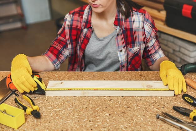 Zdjęcie bliska kaukaska młoda brązowowłosa kobieta w koszuli w kratę, szara koszulka, żółte rękawiczki pracująca w warsztacie stolarskim na drewnianym stole z różnymi narzędziami, mierząca długość paska za pomocą taśmy mierniczej