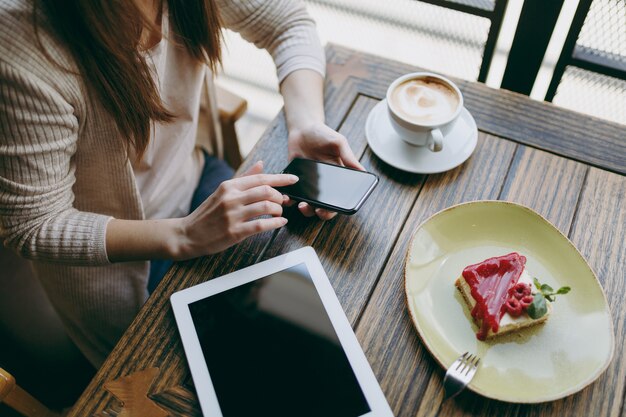 Bliska kobieta w kawiarni z komputera typu tablet pc filiżankę ciasta cappuccino zrelaksować się w restauracji w czasie wolnym. Kobieta trzyma telefon komórkowy z pustym pustym ekranem odpoczynku w kawiarni. Koncepcja stylu życia.