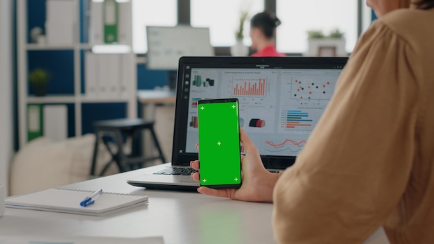 Bliska kobiety pionowo trzymając smartfon z zielonego ekranu w biurze firmy. Osoba patrząca na telefon komórkowy z na białym tle makiety tła i szablonu klucza chrominancji na wyświetlaczu.