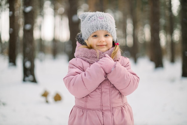 Bliska odkryty portret cute dziewczynka, na sobie różową kurtkę, czapkę i rękawiczki, pozowanie z uśmiechem do kamery na tle śniegu zima lasu.