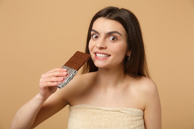 Bliska półnaga kobieta o idealnej skórze, nagi makijaż trzyma tabliczkę czekolady odizolowaną na beżowej pastelowej ścianie