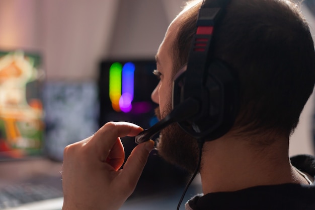 Bliska portret gracza pro esport rozmawiającego na zestawie słuchawkowym z kolegami z drużyny podczas wirtualnego turnieju gier online. Domowa sala studyjna z neonami RGB, odtwarzacz cyfrowy