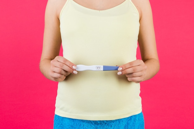 Bliska portret kobiety w ciąży w kolorowej odzieży domowej, posiadającej pozytywny wynik testu ciążowego na brzuchu na różowej powierzchni