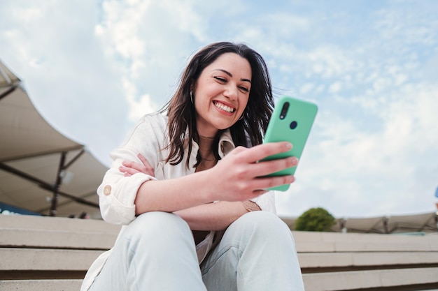 Bliska portret młodej brunetki oglądającej treści w aplikacji na telefon komórkowy siedzącej na zewnątrz Beztroska dama uśmiechająca się i dobrze się bawiąca wysyłając SMS-y za pomocą smartfona