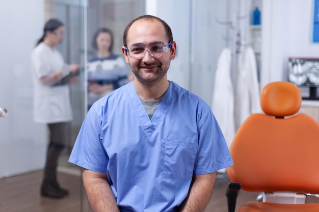 Bliska portret ortodonta w gabinecie stomatologicznym z pacjentami w tle. Stomatolog w klinice zębów professioanl uśmiechający się na sobie mundur patrząc na kamery.