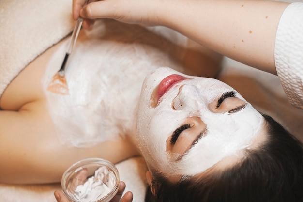 Bliska portret pięknej młodej kobiety rasy kaukaskiej o białej masce do pielęgnacji skóry organicznej na twarzy, podczas gdy jej kosmetolog nakłada aloes na szyję.