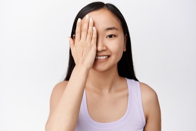 Bliska portret ślicznej azjatyckiej dziewczyny bez makijażu, przeciw jednej stronie twarzy ręką, uśmiechniętą i wyglądającą wesoło na biało na białym