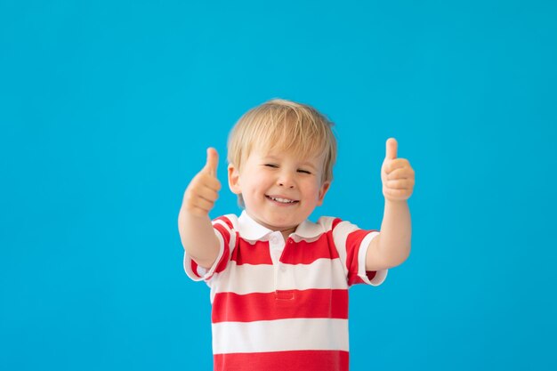 Bliska Portret Szczęśliwego Dziecka Na Sobie Koszulę W Paski Pokazując Kciuki Do Góry Przed Niebieską ścianą.