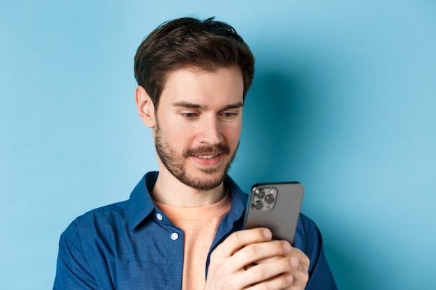 Bliska portret szczęśliwy młody człowiek czytający telefon komórkowy sreen, tworzenie sieci w smartfonie, stojąc na niebieskim tle.