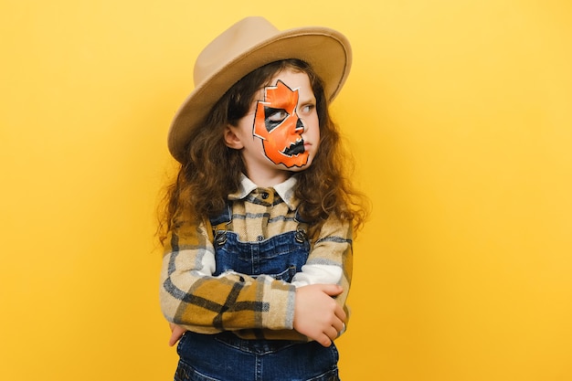 Zdjęcie bliska portret zły smutny dziecko dziewczynka z halloweenową maską do makijażu, nosi brązowy kapelusz i koszulę, skrzyżowane ręce patrząc w bok, na białym tle nad żółtą ścianą studyjną z miejsca na kopię