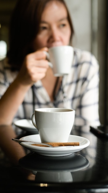 Bliska strzał Biały kubek kawy jest umieszczony na stole, młoda kobieta siedzi picia kawy w tle.