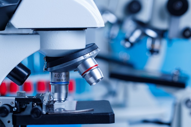 Bliska strzał naukowego mikroskopu powiększającego powiększanie monitorowania próbki szklanej płytki przed niewyraźne tło czerwone i niebieskie probówki w laboratorium szpitalnym.