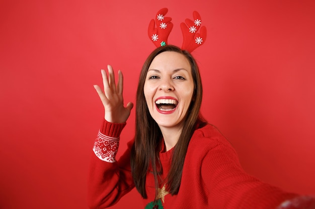 Bliska strzał selfie radosna młoda dziewczyna Santa w zabawne ozdobne rogi jelenia rosnąca ręka na białym tle na jasnym czerwonym tle. Szczęśliwego nowego roku 2019 celebracja party wakacje koncepcja. Makieta miejsca na kopię.