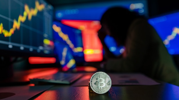 Bliska strzał srebrny bitcoin kryptowaluta wirtualny token na biurku z zdenerwowany niezadowolony niepowodzenie inwestor inwestor patrząc wykres wykres utraty wartości na ekranie monitora komputera w niewyraźne tło cień.