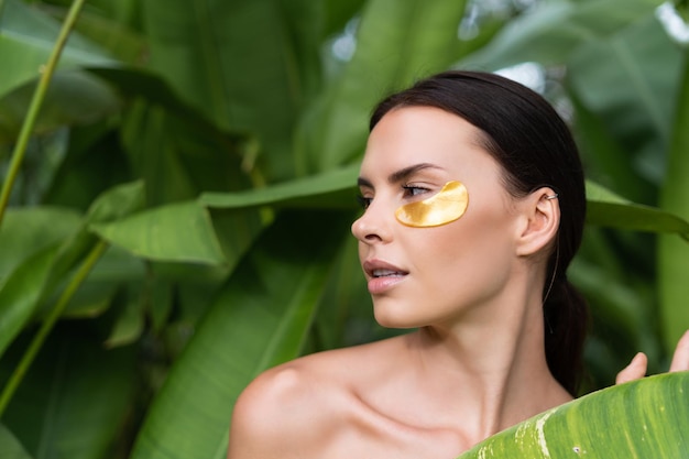 Bliski piękny portret kobiety topless z idealną skórą i naturalnym makijażem błyszczące plamy pod oczami, aby nawilżyć skórę pozującą w tropikalnych zielonych liściach na zewnątrz