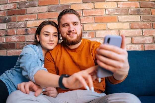 Bliski strzał portret szczęśliwej młodej pary, która ma czat online z kamerą internetową za pośrednictwem połączenia wideo na telefonie komórkowym, siedząc razem na wygodnej miękkiej sofie w przytulnym salonie. Pojęcie życia domowego.