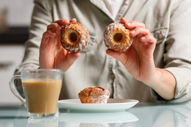 bliskie ręce trzymające i pokazujące dwa muffiny na talerzu z innym ciastem i szklanym kubkiem kawy z mlekiem stojącym na stole