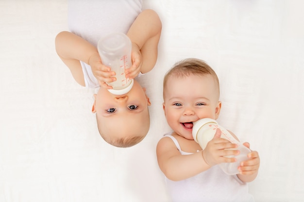 Bliźniaczy chłopiec i dziewczynka z butelką mleka na białym łóżku w domu, koncepcja żywności dla niemowląt