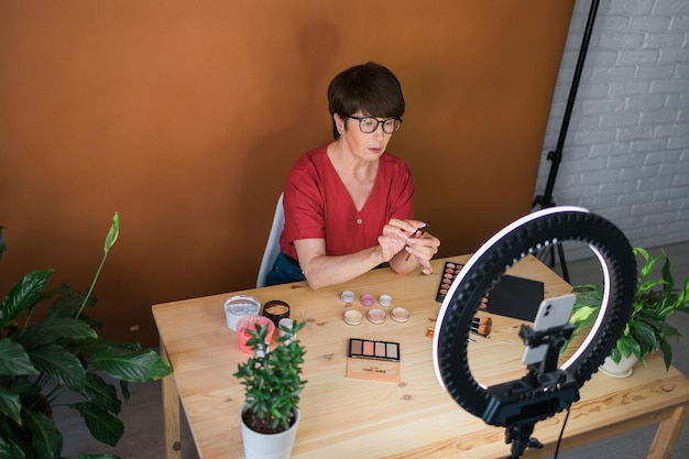 Blogerka lub influencerka zajmująca się urodą w średnim wieku z produktem do makijażu nagrywa wideo w pokoju w domu za pomocą lampy pierścieniowej i smartfona