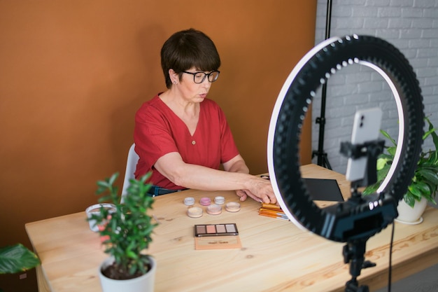 Blogerka lub influencerka zajmująca się urodą w średnim wieku z produktem do makijażu nagrywa wideo w pokoju w domu za pomocą lampy pierścieniowej i smartfona