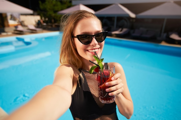 Blogerka podróżnicza w bikini robiąca selfie z koktajlem w pobliżu basenu Pov of lifestyle vlogger selfie z wakacji w luksusowym kurorcie Kobieta w stroju kąpielowym pije napoje w tropikalnym spa