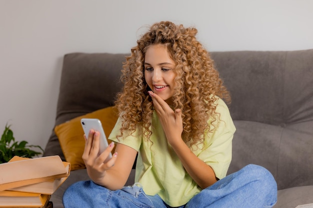 Blogerka ze słuchawkami i smartfonem w dłoni siedzi ze skrzyżowanymi nogami na szarej sofie. młoda kobieta z kręconymi włosami komunikuje się online. Zdjęcie wysokiej jakości