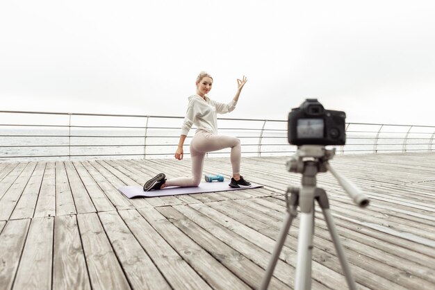 Blogowanie fitness. Młoda zdrowa kobieta sport rejestruje trening jogi w aparacie ze statywem, pokazując, jak wykonywać asany na plaży. vlogowanie