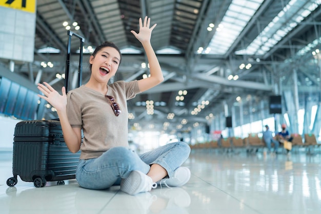 Blokada jest z biegiem czasu do podróżyszczęście azjatyckie femlae dorywczo tkanina gest machania ręką uśmiecha się, gdy siedzisz relaks na podłodze lotniska terminala z koncepcją podróży bezpieczeństwa bagażu
