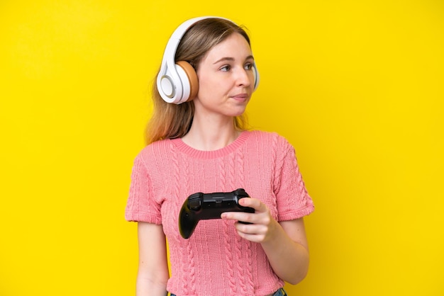 Blond angielska młoda dziewczyna bawi się kontrolerem gier wideo odizolowanym na żółtym tle, patrząc z boku