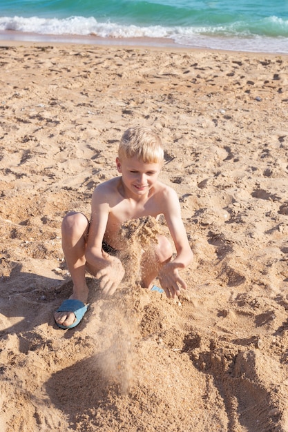 Blond chłopiec bawi się piaskiem na plaży. Letnie szczęśliwe wakacje nad morzem.
