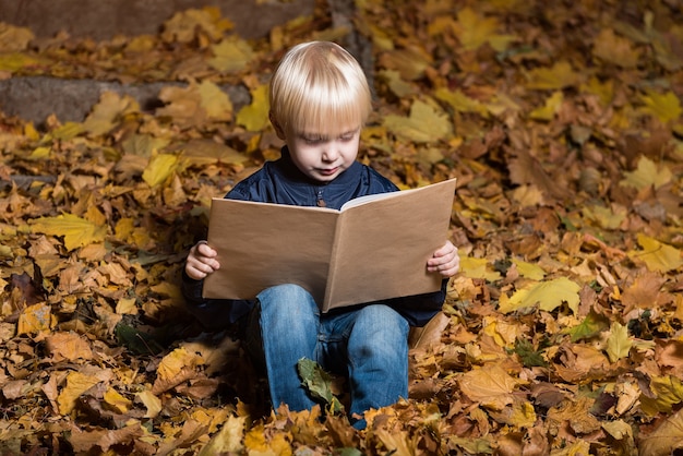 Blond chłopiec czytanie książki w lesie jesienią, siedząc na opadłych liściach. Portret.