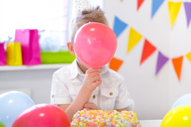 Blond chłopiec kaukaski ukrył się za czerwony balon w pobliżu urodzinowego tęczowego ciasta. Świąteczne kolorowe tło. Śmieszne przyjęcie urodzinowe