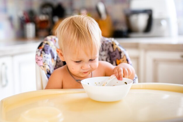 Blond dziecko w foteliku jedzenie zupy. Małe dziecko w krzesełko do jedzenia miękkich warzyw łyżką. Pierwszy stały pokarm dla dziecka w domu.