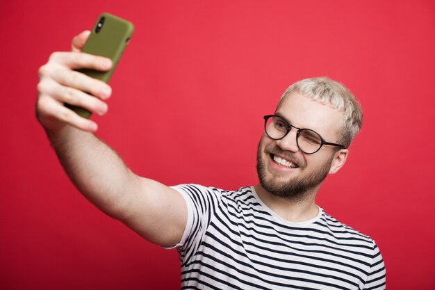 Blond kaukaski mężczyzna w okularach robi selfie na czerwonej ścianie