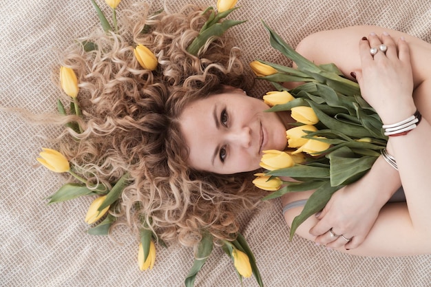 Blond kędzierzawa kobieta leży na łóżku z wiosennym bukietem żółtych tulipanów widok z góry