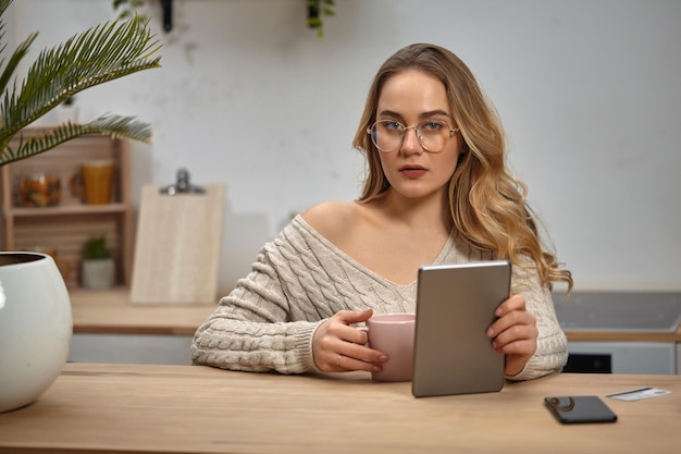 Blond kobieta blogger w okularach beżowy sweter z dzianiny trzymając tablet i różowy kubek siedzi w kuchni przy drewnianym stole z palmą w białym garnku smartfon i plastikowa karta na nim Zbliżenie