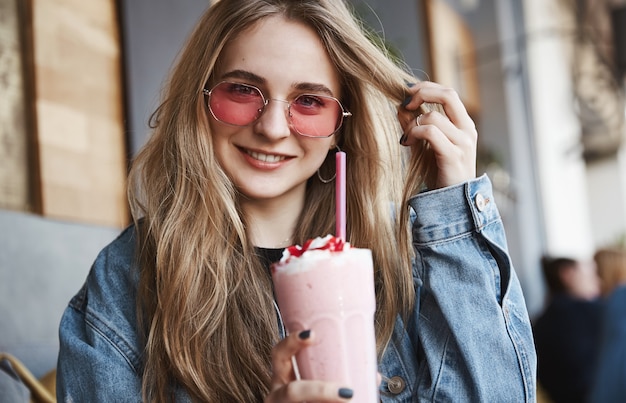 Blond kobieta w okularach przeciwsłonecznych pije koktajl truskawkowy w kawiarni