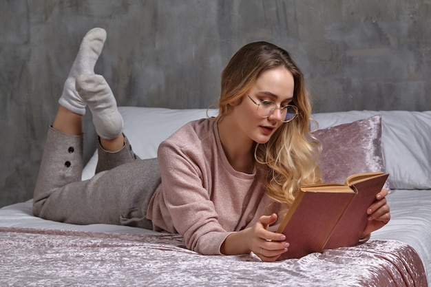 Blond kobieta w okularach ubranie na co dzień leży na łóżku i czyta książkę w sypialni uczen...