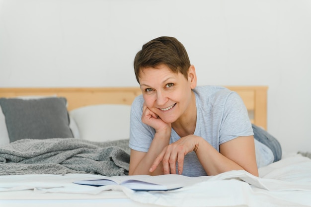 Blond kobieta w średnim wieku czyta książkę leżącą na łóżku w domu