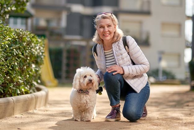 Blond kobieta w średnim wieku spacerująca z puszystym białym psem w letnim mieście.