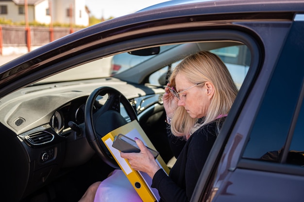 Blond kobieta w średnim wieku w okularach w samochodzie pracuje z dokumentami koncepcja technologii mobilnej