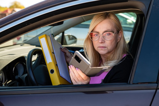 Blond kobieta w średnim wieku w okularach w samochodzie pracuje z dokumentami koncepcja technologii mobilnej