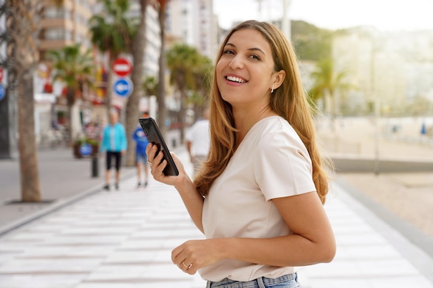 Blond Modelka Trzymająca Telefon Rozgląda Się Po Promenadzie Z Rozmytym Tłem Miasta Z Plamami Drzew I Drapaczami Chmur