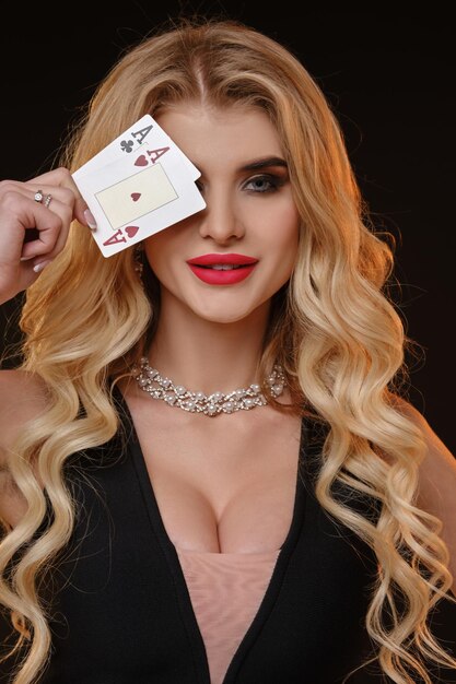 Blond modelka w czarnej sukience i naszyjniku. Uśmiechnęła się, zakryła oko dwiema kartami do gry, pozując na czarnym tle. Poker, kasyno. Zbliżenie